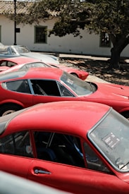 Several vintage sports cars are parked under the shade of a large tree beside a building. The cars have a classic design, with vibrant red colors dominating the scene. There's a mix of coupe and convertible models, showcasing their distinct curves and body styles.