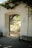 An arched doorway in a white plastered wall opens to a garden with lush greenery and trees. Sunlight filters through leaves, casting shadows on the ground. A wooden bench sits to the side, and the words 'The Pacific House' are painted on the wall.