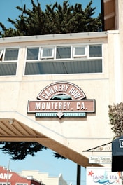 A sign reading 'Cannery Row, Monterey, CA' is displayed prominently on an overhanging structure, with additional text 'Steinbeck Plaza Two'. The background features the tops of buildings and lush green trees. A small sign for 'Louie's Kitchen' and another for 'Sand & Sea' are visible below the main sign.