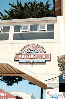 A sign reading 'Cannery Row, Monterey, CA' is displayed prominently on an overhanging structure, with additional text 'Steinbeck Plaza Two'. The background features the tops of buildings and lush green trees. A small sign for 'Louie's Kitchen' and another for 'Sand & Sea' are visible below the main sign.