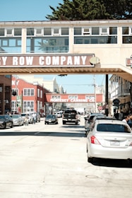 A busy street scene with several parked and moving cars under a pedestrian bridge labeled Cannery Row Company. The area features historical industrial architecture with red brick buildings and signage for the Monterey Canning Co. People are visible walking and cycling on the sidewalk.