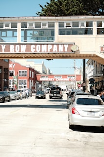 A busy street scene with several parked and moving cars under a pedestrian bridge labeled Cannery Row Company. The area features historical industrial architecture with red brick buildings and signage for the Monterey Canning Co. People are visible walking and cycling on the sidewalk.