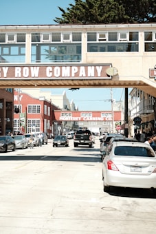 A busy street scene with several parked and moving cars under a pedestrian bridge labeled Cannery Row Company. The area features historical industrial architecture with red brick buildings and signage for the Monterey Canning Co. People are visible walking and cycling on the sidewalk.