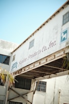 An industrial building with a white exterior, featuring a sign that reads 'Sardine Products Co. Inc.' The structure includes large windows and has visible rust on metal beams. The sky is clear and blue, indicating daylight.