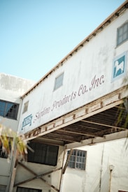 An industrial building with a white exterior, featuring a sign that reads 'Sardine Products Co. Inc.' The structure includes large windows and has visible rust on metal beams. The sky is clear and blue, indicating daylight.