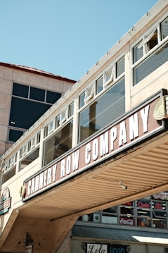 A commercial building facade with large windows and a prominent sign reading 'CANNERY ROW COMPANY'. The architecture features a beige exterior with glass panels and an overhanging structure. Underneath, there are additional storefront signs with various brand names like Coca-Cola.