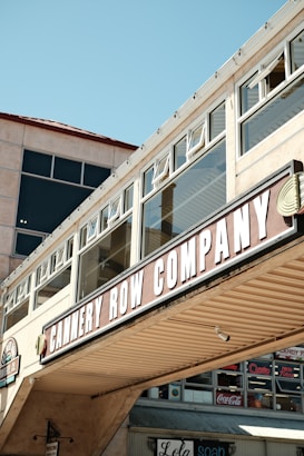A commercial building facade with large windows and a prominent sign reading 'CANNERY ROW COMPANY'. The architecture features a beige exterior with glass panels and an overhanging structure. Underneath, there are additional storefront signs with various brand names like Coca-Cola.