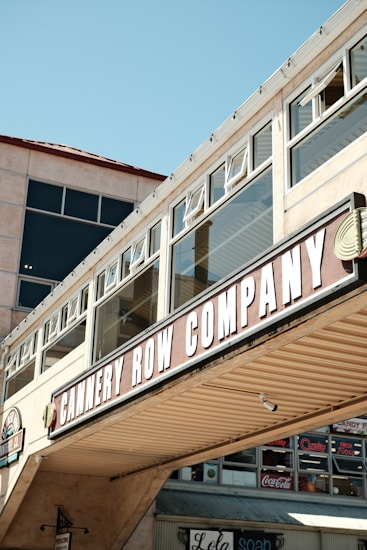 A commercial building facade with large windows and a prominent sign reading 'CANNERY ROW COMPANY'. The architecture features a beige exterior with glass panels and an overhanging structure. Underneath, there are additional storefront signs with various brand names like Coca-Cola.