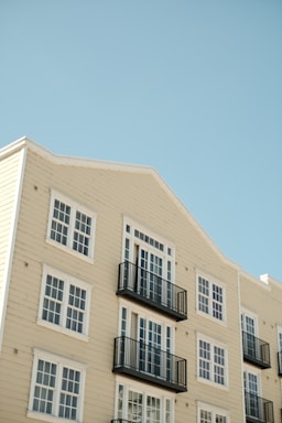 An apartment building with cream-colored siding features multiple windows and black balcony railings against a clear blue sky. The building has a clean and modern architectural style with symmetrical windows and neatly arranged balconies.