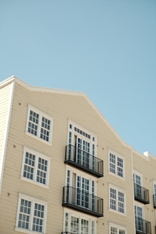 An apartment building with cream-colored siding features multiple windows and black balcony railings against a clear blue sky. The building has a clean and modern architectural style with symmetrical windows and neatly arranged balconies.