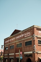 A red brick building with the words 'CANNERY ROW BREWING CO.' painted on it, along with 'EST. 2010.' The building has several large windows with black frames, and metallic barrels are arranged decoratively on the side. The structure appears industrial, and there is a blue clear sky in the background.