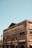 A red brick building with the words 'CANNERY ROW BREWING CO.' painted on it, along with 'EST. 2010.' The building has several large windows with black frames, and metallic barrels are arranged decoratively on the side. The structure appears industrial, and there is a blue clear sky in the background.