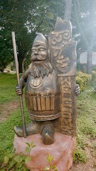 A bronze statue of a dwarf-like figure is depicted holding a vertical staff in its right hand. It has a full beard and a pointed hat. In its left hand, it holds a vertical sign with the word 'WELCOME' etched into it. Surrounding the statue is a lush green area with grass, plants, and trees in the background.