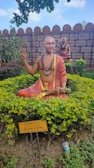 A statue of a spiritual figure sits surrounded by greenery in a garden. The statue is adorned with traditional robes, mala beads, and holds a scroll. It has a painted mark on the forehead, suggesting religious or cultural significance. In the background, another statue sits on a pedestal. The setting is enclosed by a stone wall, and a sign in front provides information about the figure, identifying it as 'MADWACHARYA' from the 13th and 14th century AD.