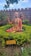 A statue of a spiritual figure sits surrounded by greenery in a garden. The statue is adorned with traditional robes, mala beads, and holds a scroll. It has a painted mark on the forehead, suggesting religious or cultural significance. In the background, another statue sits on a pedestal. The setting is enclosed by a stone wall, and a sign in front provides information about the figure, identifying it as 'MADWACHARYA' from the 13th and 14th century AD.