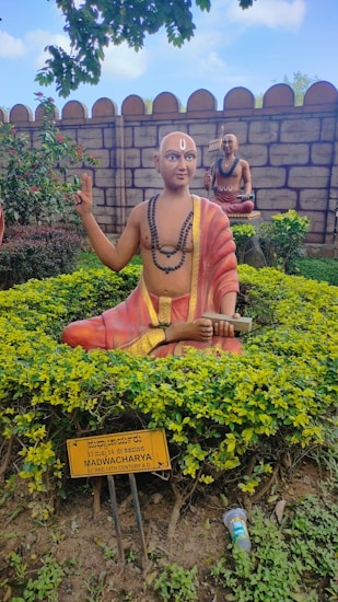 A statue of a spiritual figure sits surrounded by greenery in a garden. The statue is adorned with traditional robes, mala beads, and holds a scroll. It has a painted mark on the forehead, suggesting religious or cultural significance. In the background, another statue sits on a pedestal. The setting is enclosed by a stone wall, and a sign in front provides information about the figure, identifying it as 'MADWACHARYA' from the 13th and 14th century AD.