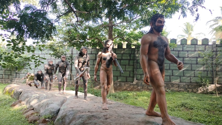 Several life-sized sculptures depict an evolutionary progression of human ancestors walking in a line on a stone path. The sculptures are set outdoors, surrounded by greenery and a brick wall in the background.