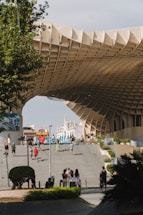 A large, modern architectural structure with a lattice-like design is prominently featured in an outdoor urban setting. People are walking and taking photos on stairs leading up to the structure. In the background, historical buildings with spires are visible against the clear sky. The scene includes greenery and a few signs with city names and directions.