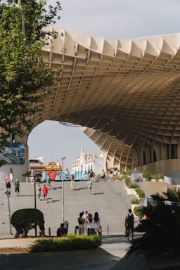 A large, modern architectural structure with a lattice-like design is prominently featured in an outdoor urban setting. People are walking and taking photos on stairs leading up to the structure. In the background, historical buildings with spires are visible against the clear sky. The scene includes greenery and a few signs with city names and directions.