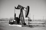 A large oil pumpjack stands in an open field with distant hills visible in the background. The machinery is metallic and weathered, surrounded by a flat barren landscape. Several pipes and power lines are visible, indicating an industrial setting.