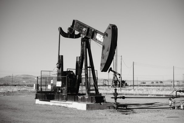 A large oil pumpjack stands in an open field with distant hills visible in the background. The machinery is metallic and weathered, surrounded by a flat barren landscape. Several pipes and power lines are visible, indicating an industrial setting.