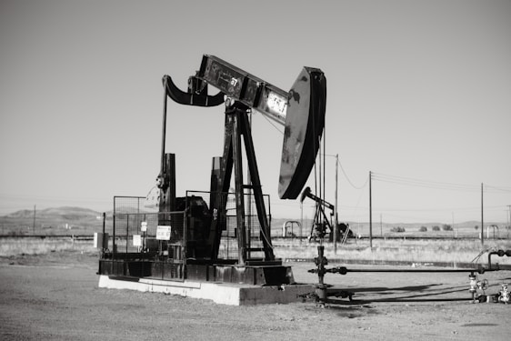 A large oil pumpjack stands in an open field with distant hills visible in the background. The machinery is metallic and weathered, surrounded by a flat barren landscape. Several pipes and power lines are visible, indicating an industrial setting.
