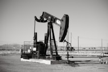 A large oil pumpjack stands in an open field with distant hills visible in the background. The machinery is metallic and weathered, surrounded by a flat barren landscape. Several pipes and power lines are visible, indicating an industrial setting.