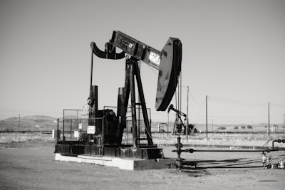 A large oil pumpjack stands in an open field with distant hills visible in the background. The machinery is metallic and weathered, surrounded by a flat barren landscape. Several pipes and power lines are visible, indicating an industrial setting.