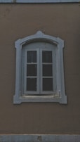 A vintage-style arched window with a worn white frame set against a textured brown wall. The window appears to have four glass panes, partly covered with dark shutters. The frame has decorative molding, adding an antique look to the overall aesthetic.