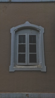A vintage-style arched window with a worn white frame set against a textured brown wall. The window appears to have four glass panes, partly covered with dark shutters. The frame has decorative molding, adding an antique look to the overall aesthetic.