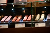 Rows of delicate, pastel-colored pastries are displayed behind a glass case in a bakery shop. The treats are arranged neatly with price tags in front, under warm lighting that highlights their details.