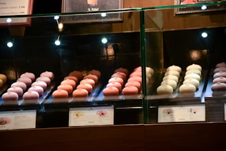 Rows of delicate, pastel-colored pastries are displayed behind a glass case in a bakery shop. The treats are arranged neatly with price tags in front, under warm lighting that highlights their details.