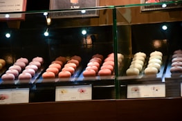 Rows of delicate, pastel-colored pastries are displayed behind a glass case in a bakery shop. The treats are arranged neatly with price tags in front, under warm lighting that highlights their details.