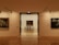 A gallery hallway with warm lighting, leading to a room featuring a large framed painting. Several black chairs are lined up in front of the painting. The gallery walls display other artworks, and the space has a minimalist and elegant design.