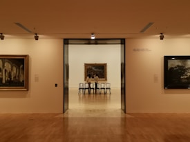 A gallery hallway with warm lighting, leading to a room featuring a large framed painting. Several black chairs are lined up in front of the painting. The gallery walls display other artworks, and the space has a minimalist and elegant design.