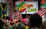 A vibrant outdoor bar setup with people serving and enjoying drinks. The signage prominently displays the text 'Rum Love' with a colorful anchor symbol. Flags and banners, including a Jamaican flag and 'Red Stripe' branding, hang prominently in the background. Decorative elements like inflatable parrots add to the festive atmosphere.