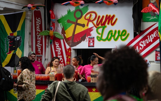 A vibrant outdoor bar setup with people serving and enjoying drinks. The signage prominently displays the text 'Rum Love' with a colorful anchor symbol. Flags and banners, including a Jamaican flag and 'Red Stripe' branding, hang prominently in the background. Decorative elements like inflatable parrots add to the festive atmosphere.