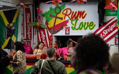 A vibrant outdoor bar setup with people serving and enjoying drinks. The signage prominently displays the text 'Rum Love' with a colorful anchor symbol. Flags and banners, including a Jamaican flag and 'Red Stripe' branding, hang prominently in the background. Decorative elements like inflatable parrots add to the festive atmosphere.
