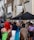 A diverse crowd of people gathers outside on a street lined with urban buildings. There is a sign for Notting Hill Community Church on one of the buildings and tents are set up nearby. The walls are adorned with graffiti and some floral decorations.
