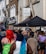 A diverse crowd of people gathers outside on a street lined with urban buildings. There is a sign for Notting Hill Community Church on one of the buildings and tents are set up nearby. The walls are adorned with graffiti and some floral decorations.