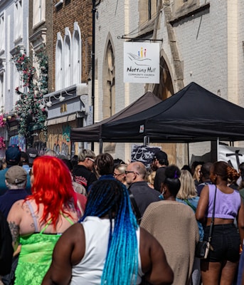 A diverse crowd of people gathers outside on a street lined with urban buildings. There is a sign for Notting Hill Community Church on one of the buildings and tents are set up nearby. The walls are adorned with graffiti and some floral decorations.