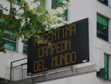A digital billboard mounted on a building displays the message 'Argentina Campeon del Mundo' in bright yellow-orange text. Trees with green leaves partly cover the top left edge of the sign, and windows of the building are visible in the background.