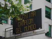 A digital billboard mounted on a building displays the message 'Argentina Campeon del Mundo' in bright yellow-orange text. Trees with green leaves partly cover the top left edge of the sign, and windows of the building are visible in the background.