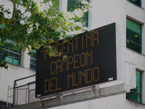 A digital billboard mounted on a building displays the message 'Argentina Campeon del Mundo' in bright yellow-orange text. Trees with green leaves partly cover the top left edge of the sign, and windows of the building are visible in the background.
