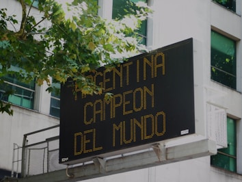 A digital billboard mounted on a building displays the message 'Argentina Campeon del Mundo' in bright yellow-orange text. Trees with green leaves partly cover the top left edge of the sign, and windows of the building are visible in the background.