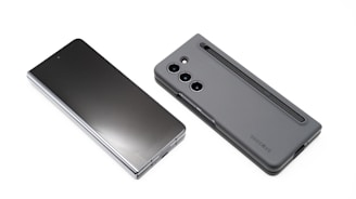 Two modern smartphones are displayed on a white background. One phone lies flat with its screen facing upward, showcasing a sleek, reflective surface. The other phone, seen from the back, features a matte case with a camera module and a stylus inserted into a slot.