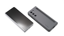 Two modern smartphones are displayed on a white background. One phone lies flat with its screen facing upward, showcasing a sleek, reflective surface. The other phone, seen from the back, features a matte case with a camera module and a stylus inserted into a slot.