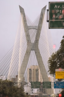 A modern cable-stayed bridge with a prominent X-shaped design dominates the view. Several road signs with text in Portuguese are visible in the foreground, alongside green foliage. The sky is overcast, and there are buildings partially obscured by the bridge's supports.