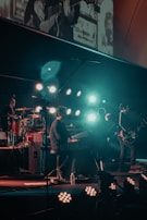 A band performing on stage with various musical instruments, including a keyboard, drum set, and electric guitar. The stage is illuminated by multiple bright lights, creating a vibrant atmosphere. Band members are engaged in playing their instruments, contributing to a dynamic live music setting.