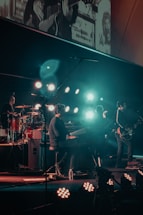 A band performing on stage with various musical instruments, including a keyboard, drum set, and electric guitar. The stage is illuminated by multiple bright lights, creating a vibrant atmosphere. Band members are engaged in playing their instruments, contributing to a dynamic live music setting.